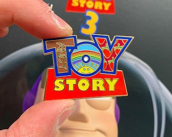 Könnte beinhalten: Eine goldfarbene Anstecknadel mit den Worten "Toy Story" in roten und blauen Buchstaben. Der "Toy"-Teil des Logos besteht aus einer regenbogenfarbenen Schallplatte mit einer Smiley-Sonne in der Mitte. Der Name "Andy" ist seitlich auf der Schallplatte in braunen Buchstaben geschrieben.