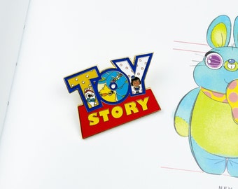 May include: A gold-colored enamel pin featuring the Toy Story logo with Woody, Buzz Lightyear, and Jessie peeking out from behind the letters. The pin is red, yellow, and blue.