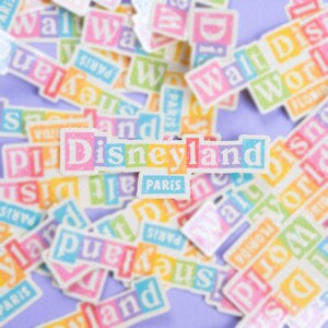 May include: A collection of colorful stickers with the words "Disneyland Paris" in a rainbow color scheme. The stickers are arranged in a random pattern on a light purple background.