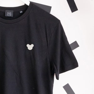 Könnte beinhalten: Schwarzes T-Shirt mit einem kleinen, weißen, gestickten Mickey-Maus-Kopf auf der linken Brust.