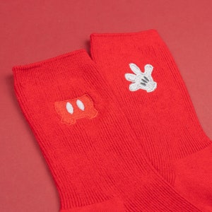 以下が含まれることがあります： 赤い靴下2足に、白いミッキーマウスの刺繍が施されています。片方の靴下にはミッキーの象徴的な手袋が、もう片方には耳が刺繍されています。