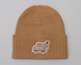 Puede incluir: Un gorro de punto acanalado color canela con un dobladillo. Un parche bordado en el dobladillo muestra una grúa marrón con el texto "TOW MATER TOWING & SALVAGE" en letras de color marrón oscuro, delineado en blanco sobre la tela canela.