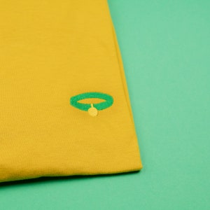 May include: A mustard yellow t-shirt with a green embroidered dog collar and a yellow bell charm.