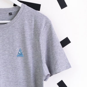 Może przedstawiać: Szary t-shirt z małym niebieskim nadrukiem w kształcie kapelusza czarodzieja z przodu. Kapelusz ma półksiężyc i gwiazdy.
