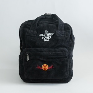 May include: Black corduroy backpack featuring the text "The Hollywood Tower Hotel" in white. The backpack has a front pocket with a logo and a top handle. The bag has a zip closure and adjustable straps.