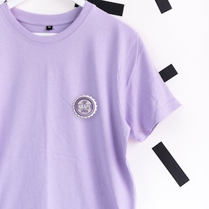 Puede incluir: Una camiseta de color lila claro con cuello redondo y mangas cortas. Un emblema plateado y morado con las palabras "GRAPE SODA" está en el pecho izquierdo. La camiseta está colgada de una percha blanca.