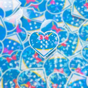 May include: A blue heart-shaped sticker with a white outline and a pink bow. The sticker is covered in iridescent glitter and has a white background.