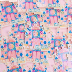 Könnte beinhalten: Ein rosa und blauer Aufkleber mit einer Cartoon-Illustration der Small World-Fahrt in Disneyland. Der Aufkleber zeigt ein Schloss mit einem Uhrturm und dem Text "Small World" in einer verspielten Schrift.
