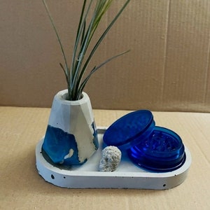 May include: A white concrete tray with a small white concrete planter with a blue and white design. The planter has a green plant inside. The tray also has a blue grinder and a small white rock.