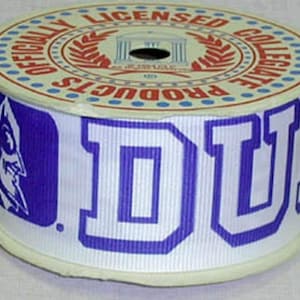 May include: A spool of white ribbon with the word "DUKE" in large, blue block letters. The ribbon also features the Duke Blue Devils logo and the text "OFFICIALLY LICENSED COLLEGIATE PRODUCTS" in red and blue.