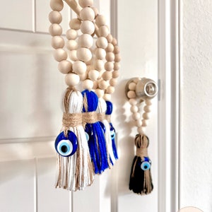 May include: Three wooden bead garlands with blue and white tassel charms featuring evil eye beads. The garlands are hanging on a white door.