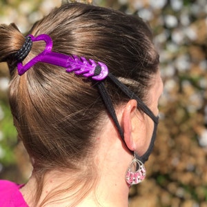 Pode incluir: Um grampo de cabelo de plástico roxo com um design em forma de coração é usado para segurar um rabo de cavalo no lugar. O grampo está preso a uma faixa elástica preta que está segurando uma máscara facial no lugar.