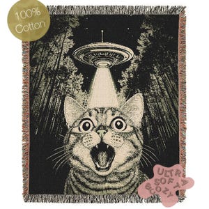 May include: A black and white woven tapestry featuring a cat with wide eyes and an open mouth looking up at a UFO in the sky. The UFO is emitting a beam of light. The background is a dark forest with trees and stars. The tapestry has a fringe border and a label that says "Ultra Soft Cozy".