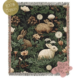 Puede incluir: Una manta tejida con un fondo oscuro con conejos, flores y follaje. La manta tiene un borde con flecos y el texto "100% Cotton" es visible. La manta está etiquetada como "Ultra Soft & Cozy".