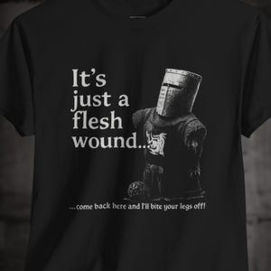 May include: Black t-shirt with a humorous illustration of a medieval knight. The text says "It's just a flesh wound... come back here and I'll bite your legs off!" The knight is in full armour.