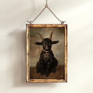 May include: A framed portrait of a black goat with horns and a pentagram necklace. The artwork is in a gold-colored frame and is suspended by a chain. The goat is sitting and has a serious expression.