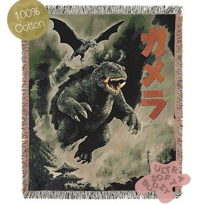 Puede incluir: Una manta de algodón tejido con un monstruo y una criatura voladora en una escena de batalla. La manta tiene un borde con flecos y el texto "100% Cotton" es visible. También está presente el texto japonés "ガメラ".