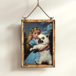 May include: Framed artwork depicting a young girl in a blue dress holding a snarling white dog. The painting has a vintage aesthetic, with a gold-colored frame and a chain for hanging. The background features a cloudy sky and trees.