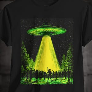 May include: Black t-shirt featuring a green and yellow graphic of a UFO beaming light onto a crowd of people in a forest. The design includes a starry night sky and silhouettes of trees.