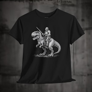 May include: Black t-shirt featuring a detailed black and white illustration of a knight in full armor riding a Tyrannosaurus Rex, holding a sword. The design is centered on the shirt, showcasing a fantasy theme.