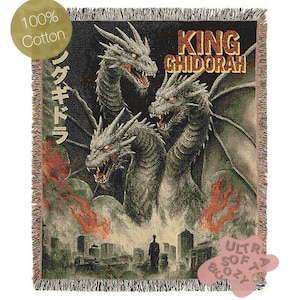 May include: A woven cotton throw blanket featuring a three-headed dragon illustration with the text "KING GHIDORAH" and Japanese text. The blanket has a fringed edge and a cityscape at the bottom. The blanket is labeled "100% Cotton" and "Ultra Soft & Cozy".