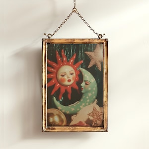 May include: A framed celestial art piece featuring a red sun with a face, a green crescent moon with stars, and a gold-colored frame. The artwork is suspended by a chain.