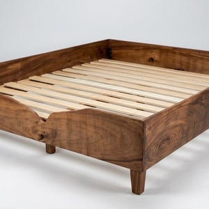Pode incluir: Uma estrutura de cama de perfil baixo construída em madeira castanha escura com um padrão de grão natural visível. A estrutura apresenta um recorte curvo na calha frontal e é suportada por pernas curtas e cónicas. Ripas de madeira clara visíveis.