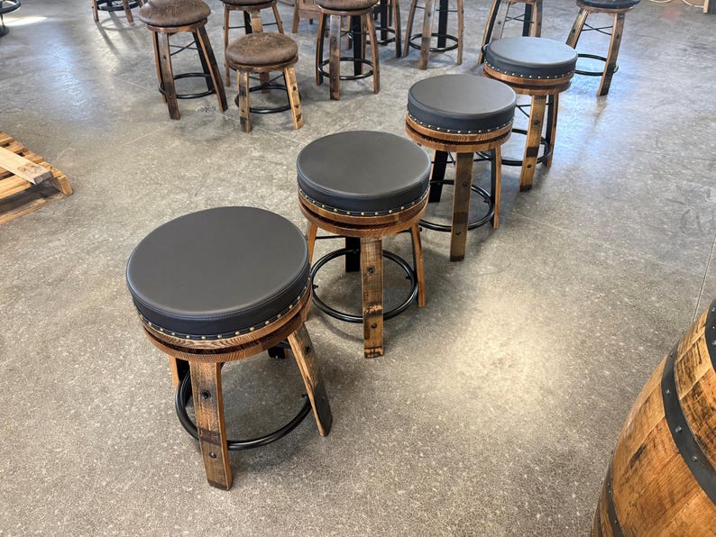 May include: A collection of bar stools featuring circular black padded seats with silver nailhead trim. The bases are constructed from curved wooden barrel staves with black metal circular footrests, providing a sturdy structure for each stool.