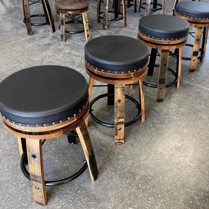 May include: A set of bar stools featuring round black padded seats with silver nailhead trim. The base of each stool is constructed from curved wooden barrel staves with a natural finish, supported by a circular black metal footrest ring.