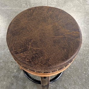 May include: A round stool featuring a dark brown, textured seat with a distressed leather-like appearance. The seat is accented with a row of small, metallic nailhead studs along the edge, supported by a wooden frame and a black circular metal footrest.