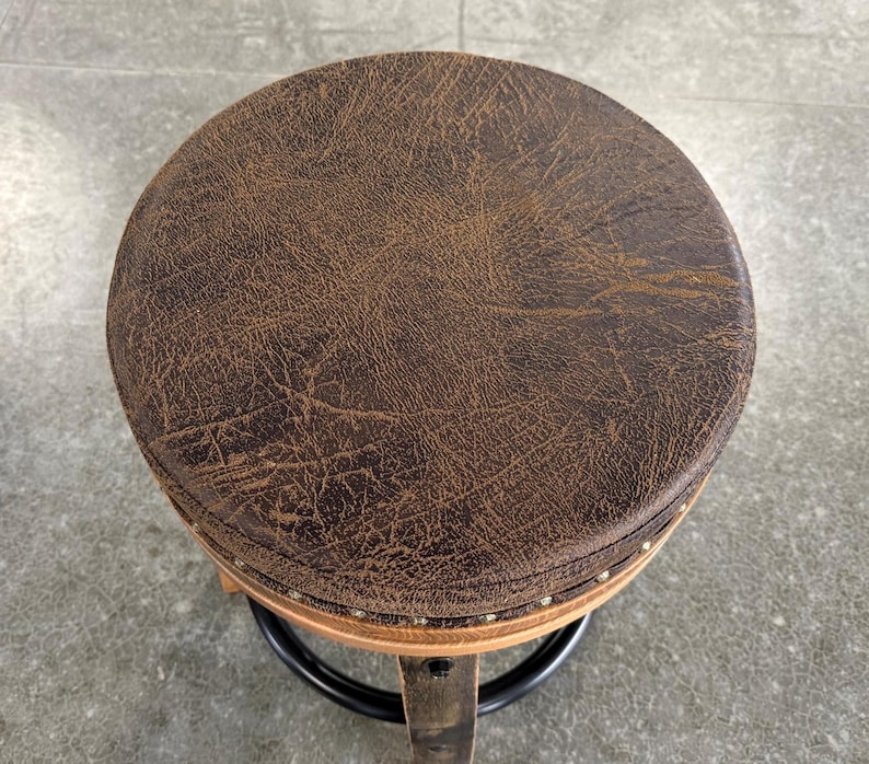 May include: A round stool featuring a dark brown, textured seat with a distressed leather-like appearance. The seat is accented with a row of small, metallic nailhead studs along the edge, supported by a wooden frame and a black circular metal footrest.