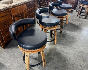 May include: A set of swivel bar stools featuring round black leather seats with silver nailhead trim, curved black backrests, and light brown wooden barrel-style legs reinforced with a circular black metal footrest ring for stability.