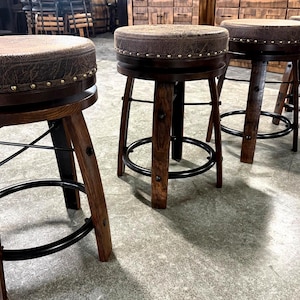 May include: A set of four round bar stools featuring dark brown wooden legs, black metal circular footrests, and swivel seats upholstered in a textured brown material. Each seat is accented with a row of metallic nailhead trim along the edge.