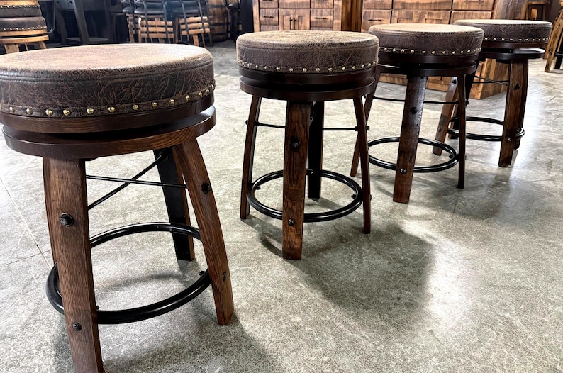 May include: A set of four round bar stools featuring dark brown wooden legs, black metal circular footrests, and swivel seats upholstered in a textured brown material. Each seat is accented with a row of metallic nailhead trim along the edge.