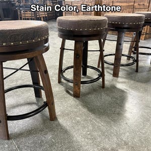 May include: Four swivel bar stools with dark brown wooden legs, black metal circular footrests, and round seats upholstered in a textured earthtone fabric. The seat edges are accented with a row of silver nailhead trim. Text reads: Stain Color, Earthtone.