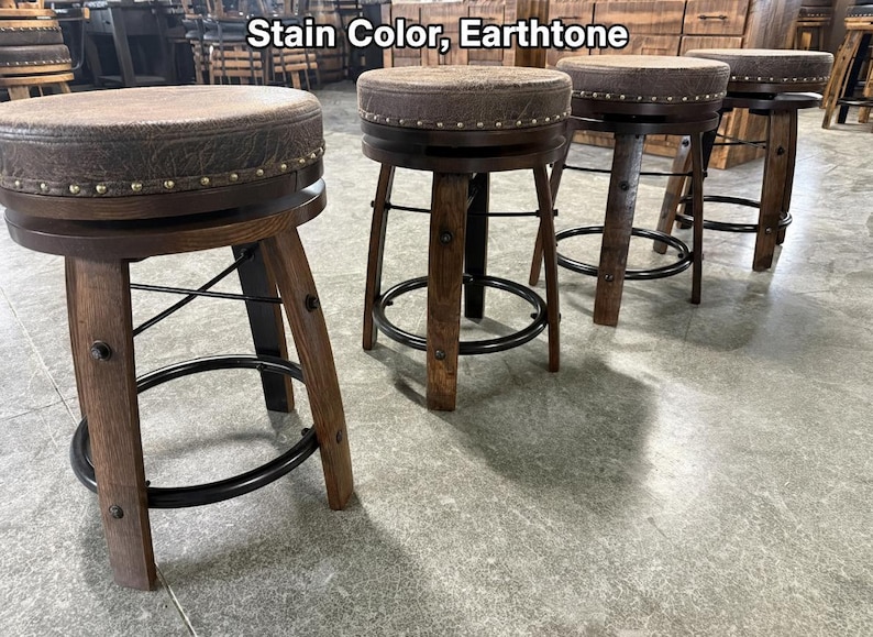 May include: Four swivel bar stools with dark brown wooden legs, black metal circular footrests, and round seats upholstered in a textured earthtone fabric. The seat edges are accented with a row of silver nailhead trim. Text reads: Stain Color, Earthtone.