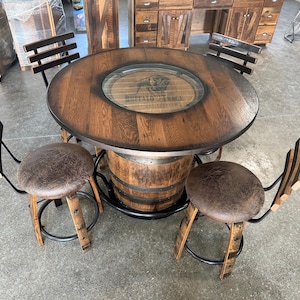 May include: A rustic, round wooden table with a glass insert, supported by a barrel base. The table is surrounded by four matching bar stools with brown leather seats and black metal frames. The table has the text "BUFFALO TRACE".