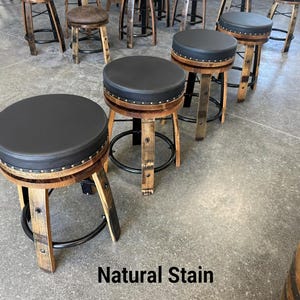 May include: A set of bar stools constructed from reclaimed wooden barrel staves with natural wood grain finishes. Each stool features a circular black padded seat, a black metal footrest ring, and decorative metal studs along the seat edge. Natural Stain.