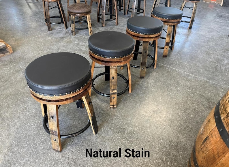 May include: A set of bar stools constructed from reclaimed wooden barrel staves with natural wood grain finishes. Each stool features a circular black padded seat, a black metal footrest ring, and decorative metal studs along the seat edge. Natural Stain.