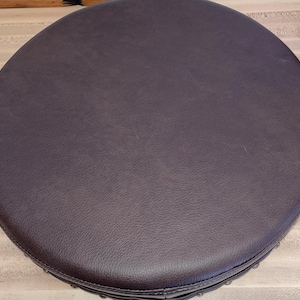 May include: A circular stool seat featuring a dark brown, textured faux leather surface. The top is perfectly round with a smooth, consistent finish across the entire area, showcasing a subtle grain pattern typical of leather upholstery materials.
