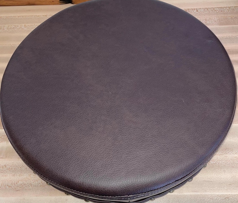 May include: A circular stool seat featuring a dark brown, textured faux leather surface. The top is perfectly round with a smooth, consistent finish across the entire area, showcasing a subtle grain pattern typical of leather upholstery materials.