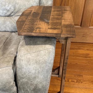 May include: A wooden side table with a dark brown finish, designed to fit over the arm of a sofa or chair. The table has a rectangular top and a supporting frame. The table is made of wood and is a practical addition to any living room.