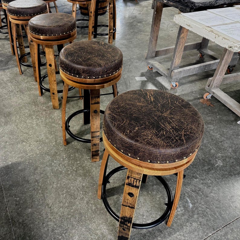 May include: A set of bar stools featuring round, dark brown, distressed leather-look seats with silver nailhead trim. The stools have curved wooden legs constructed from barrel staves and a circular black metal footrest ring at the base.