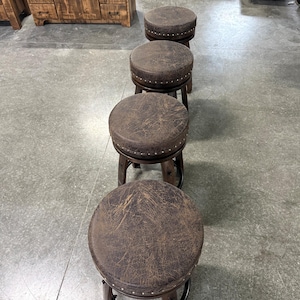 May include: A row of four round swivel stools with dark brown distressed leather-effect seats. Each seat features a circular top with a band of small silver-toned metal nailhead trim around the edge, supported by a dark wooden base structure.