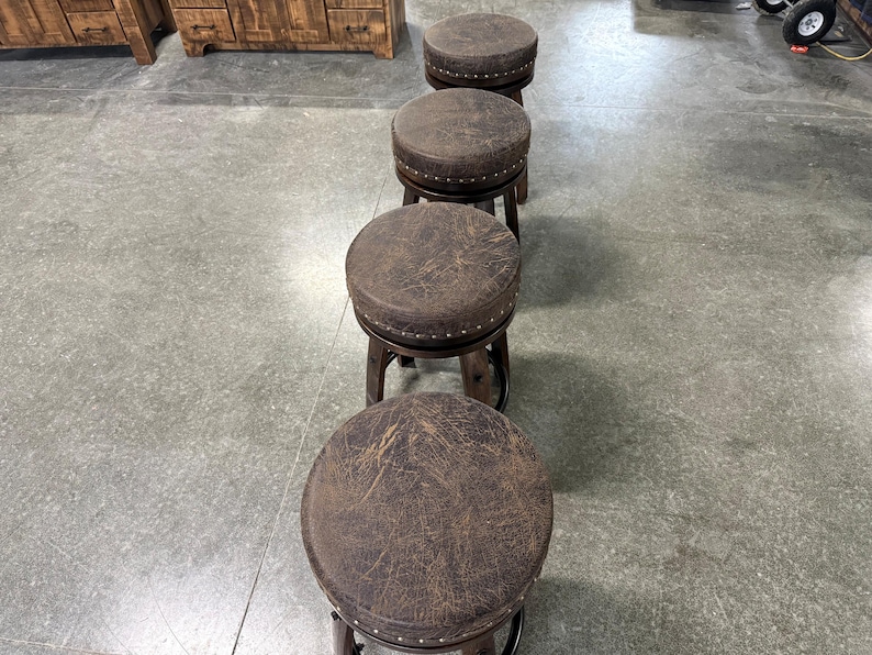May include: A row of four round swivel stools with dark brown distressed leather-effect seats. Each seat features a circular top with a band of small silver-toned metal nailhead trim around the edge, supported by a dark wooden base structure.