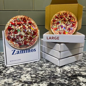 May include: Two pizzas with toppings, one in a box labeled "Zaminos" and the other in an open box labeled "LARGE". The boxes are white with blue text. The pizzas have a variety of toppings.