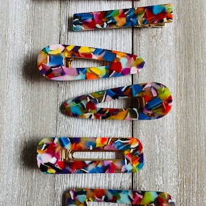 May include: Set of six colorful hair clips with a marbled pattern. The clips are made of plastic and have a gold metal spring.