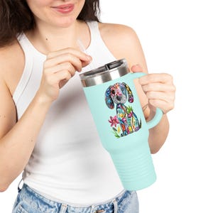 May include: A light blue travel mug with a handle and a silver lid. The mug features a colourful, stylised illustration of a dog with floral and geometric patterns. A clear straw is inserted into the lid.