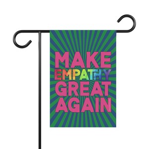 May include: A garden flag with the words "MAKE EMPATHY GREAT AGAIN" in pink and rainbow lettering. The flag has a green and blue striped background and is mounted on a black metal stand.