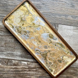 Puede incluir: Una bandeja rectangular de madera con una incrustación de resina marmoleada dorada y plateada. La bandeja tiene un borde redondeado y es perfecta para servir o exhibir artículos.